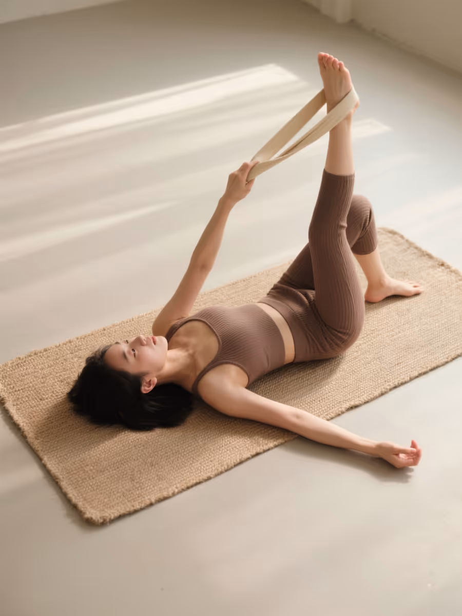 Woman lying on a mat stretching her leg upward using a yoga strap in a bright room.